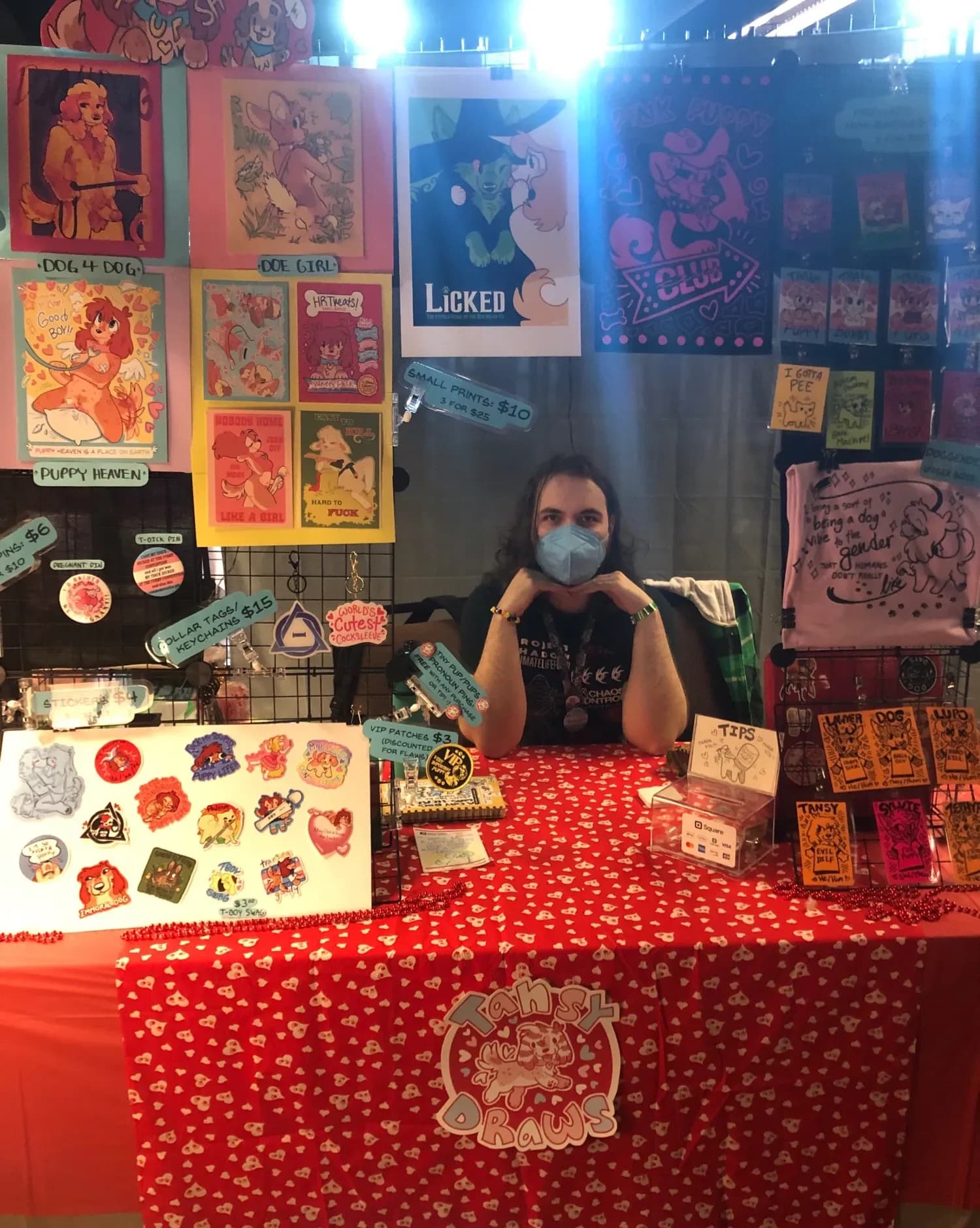 An artist alley table at Anthro New England 2026, with prints titled Puppy Heaven, ECC Girl, ECC Club, and Licked hung as a backdrop, alongside enamel pins, sticker sheets, and merchandise displayed on a red tablecloth.