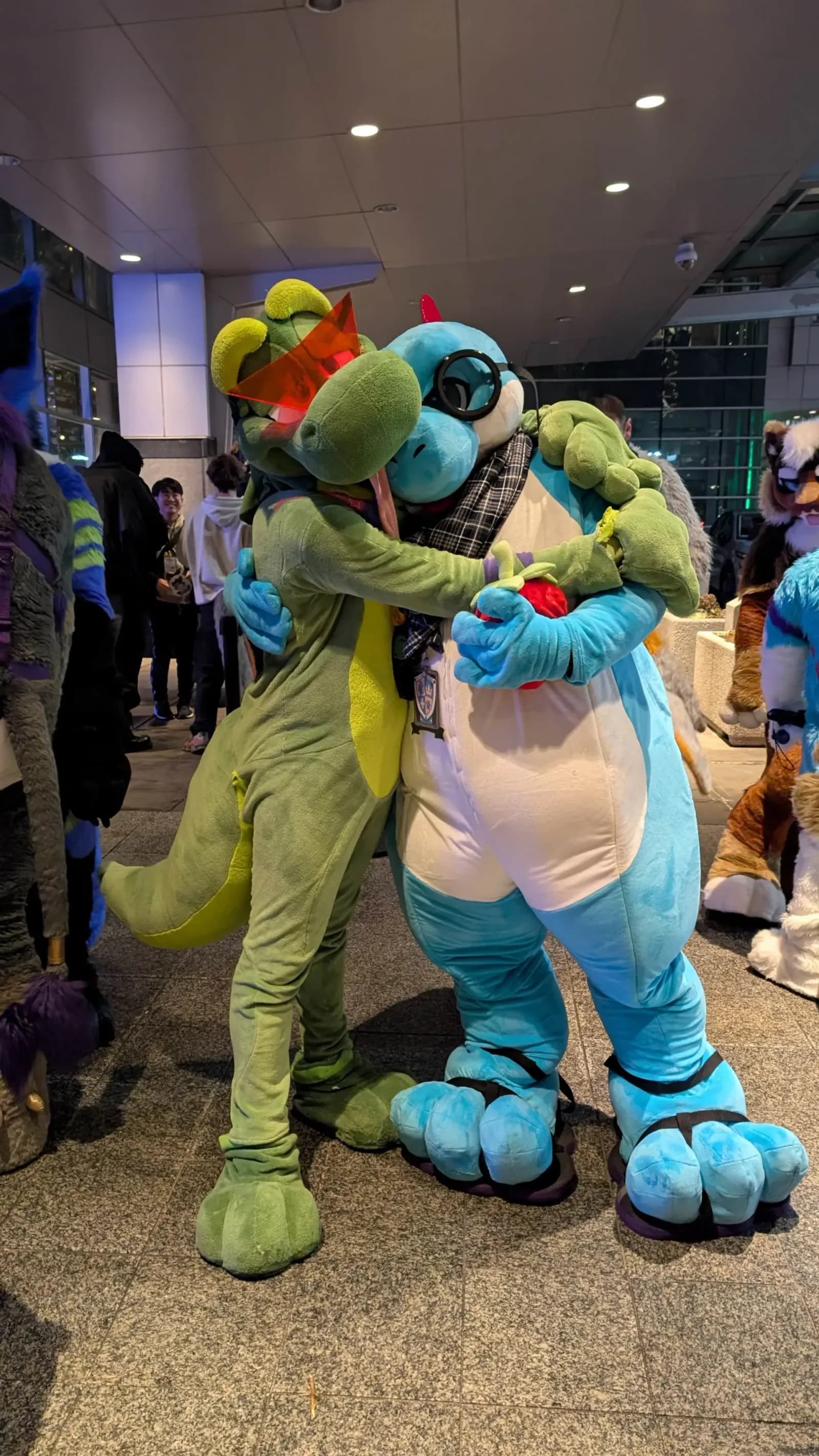 Two fursuit dinosaur characters (one green, one teal) hugging in the Westin Boston Seaport District atrium at Anthro New England 2026.