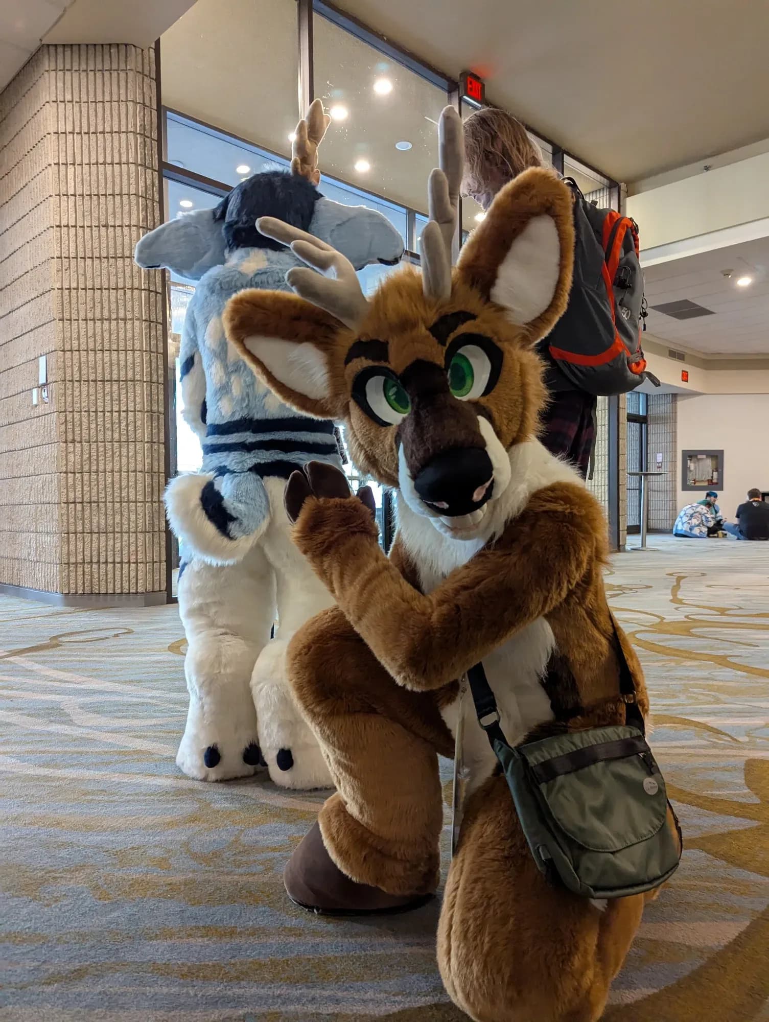 Two fursuiters in the Furnal Equinox venue lobby: a brown-and-white reindeer-style character in the foreground waving, with a black-and-white striped suit standing behind.