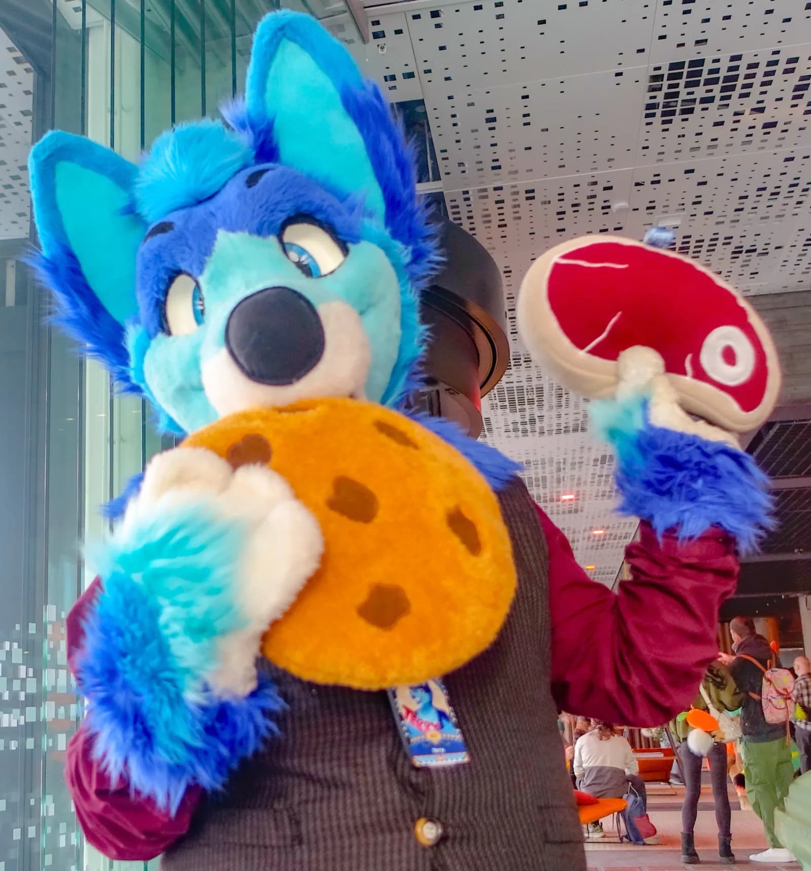 A blue-husky-style fursuiter at NordicFuzzCon 2026 holding two oversized plush props (a giant cookie and a steak) inside Malmö Mässan.