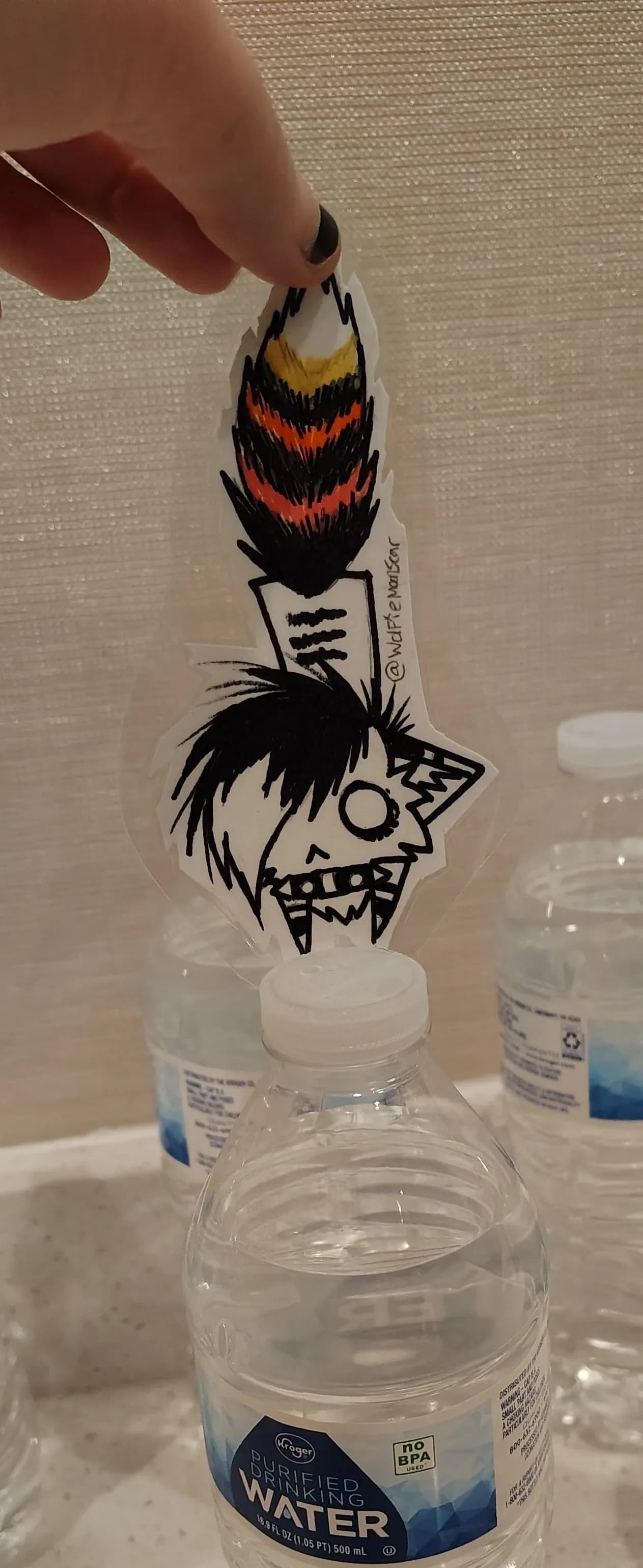 A hand-held clear-vinyl sticker featuring a feather-headdress character mark, photographed against the convention-hotel backdrop with branded water bottles in the foreground.