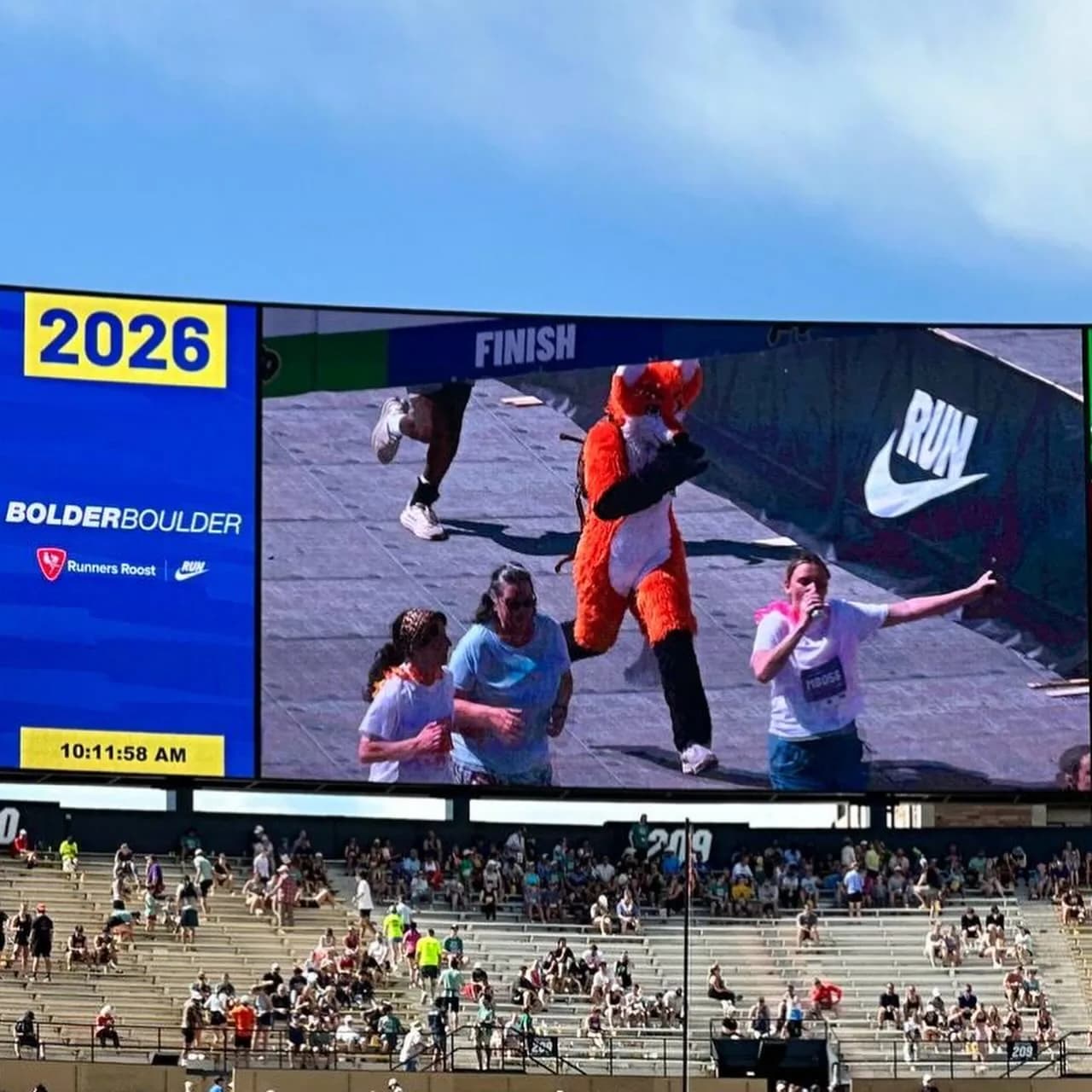 The Folsom Field jumbotron at BolderBoulder 2026 showing Blaze the Foxx (a bright orange fox fursuit) running across a finish-line camera between two other runners. The score-bug overlay reads '2026 BOLDERBoulder · Runners Roost · Nike · 10:11:58 AM.'