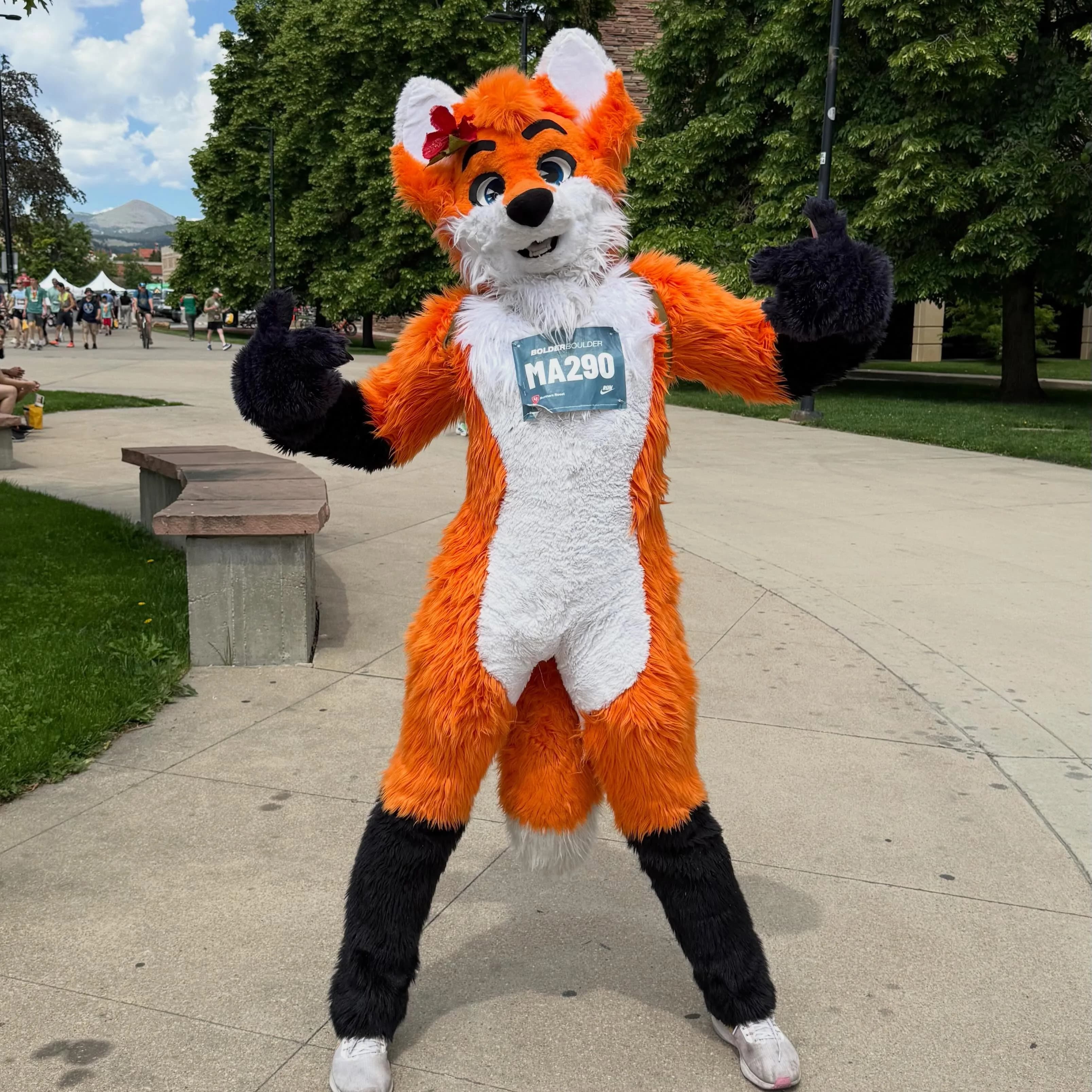 Blaze the Foxx in a relaxed pose with one paw on his hip and one raised, on a Boulder sidewalk near a tree-lined park, BolderBoulder festival tents visible behind him.