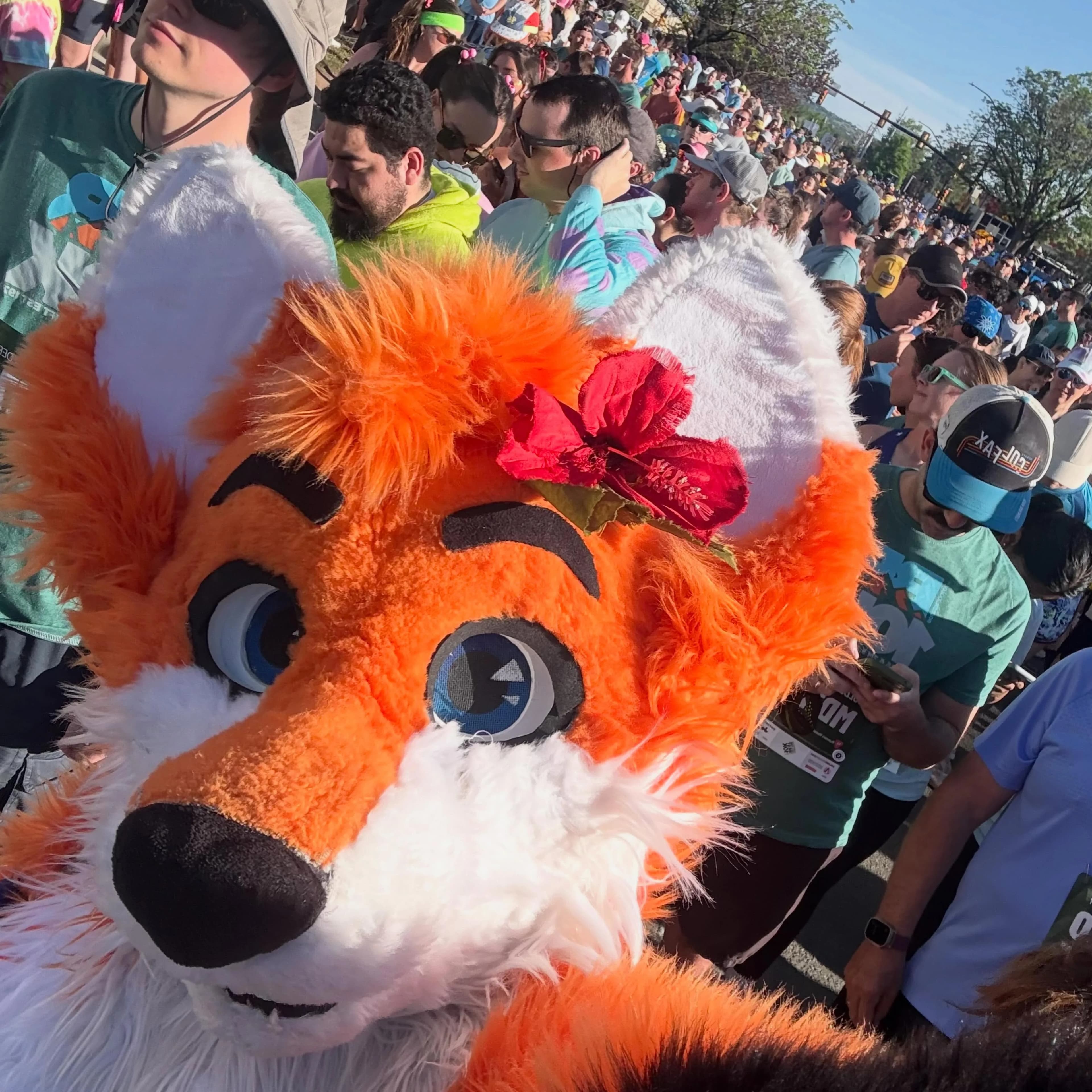 Selfie-angle close-up of Blaze the Foxx in a dense post-race crowd at golden hour, with the hibiscus flower still tucked behind his ear and his head turned slightly to the camera.