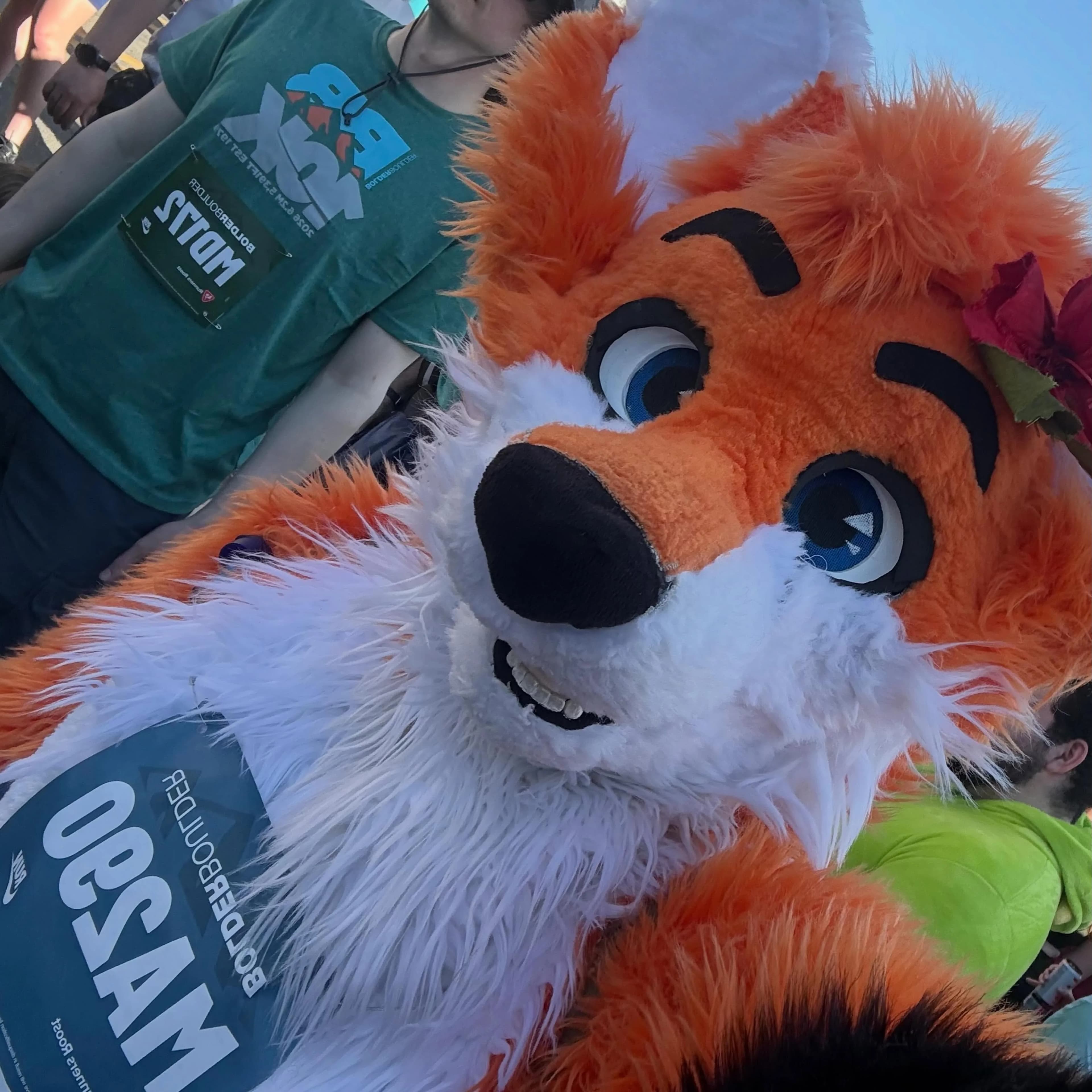 Tilted selfie of Blaze the Foxx with a finisher in a green BolderBoulder shirt, the MA290 bib lower-left and the fox's hibiscus flower still visible behind his ear.